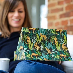 May include: A woman is holding a gift bag with a green and yellow jungle print featuring leopards. The bag is folded in half and the woman is smiling.
