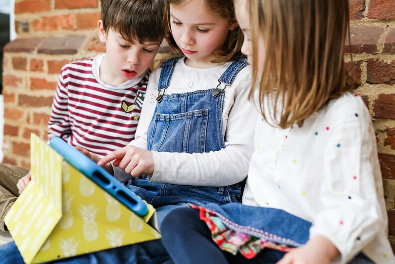 May include: Three children are sitting on a brick step, looking at a tablet computer in a yellow pineapple-patterned case. The tablet is blue and has a white case.