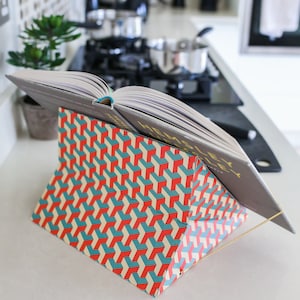 K&ouml;nnte beinhalten: Ein Kochbuch mit dem Titel "Hemsley + Hemsley" ist auf einem geometrisch gemusterten Buchst&auml;nder aufgeschlagen. Der St&auml;nder besteht aus Pappe und hat einen goldenen Metalldrahtrahmen.