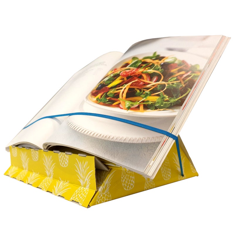 K&ouml;nnte beinhalten: Ein gelber Papp-Kochbuchst&auml;nder mit Ananas-Muster. Ein Kochbuch mit einem Rezept f&uuml;r einen Salat ist auf dem St&auml;nder aufgestellt. Ein blaues Gummiband h&auml;lt das Kochbuch geschlossen.