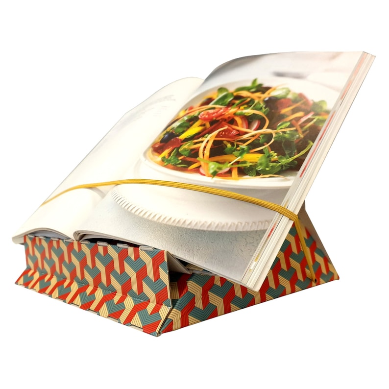 K&ouml;nnte beinhalten: Ein bunter, geometrisch gemusterter Buchst&auml;nder mit einem aufgeschlagenen Kochbuch darauf. Das Kochbuch wird mit einem gelben Gummiband offen gehalten und zeigt ein Foto eines Salats.