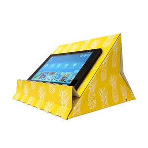 May include: A yellow cardboard tablet stand with a pineapple pattern. The stand is folded up and a tablet is resting on top of it.
