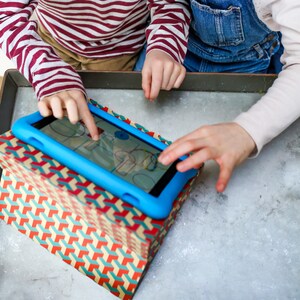 May include: Two children are using a blue tablet computer with a colorful screen. The tablet is resting on a box with a geometric pattern.