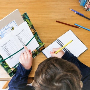 K&ouml;nnte beinhalten: Ein Kind sitzt an einem Holztisch mit einem gr&uuml;n-braunen Dschungel-Ordner, einem Notizbuch und einem Bleistift. Der Ordner hat ein gr&uuml;nes Gummiband und enth&auml;lt ein wei&szlig;es Blatt Papier mit den Worten "Spellings 22-03-19" und einer Wortliste. Das Kind schreibt in ein Notizbuch mit einem gelben Bleistift.