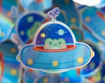 May include: A holographic sticker of a frog riding in a blue spaceship with a starry night sky background.