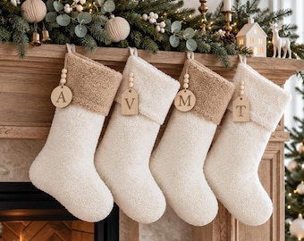 以下が含まれることがあります： クリーム色のシェルパ素材のクリスマスストッキング4足が、ライトブラウンの折り返し付きで一列に並んで吊るされています。各ストッキングには、小さな木製ビーズの紐で取り付けられた、A、V、M、Tのいずれかの文字が刻まれた木製のタグが付いており、柔らかいフリースのような質感です。