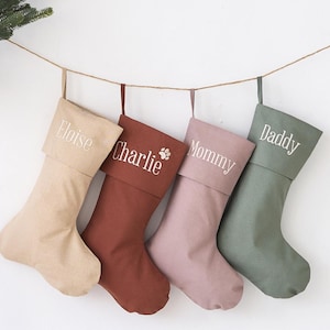 May include: Four Christmas stockings in beige, brown, pink, and green hang from a string. Each stocking is personalized with a name in white script: Eloise, Charlie, Mommy, and Daddy. A green wreath and wooden ladder are in the background.