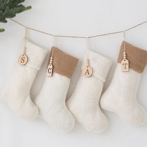 May include: Four cream-coloured Christmas stockings with brown cuffs hang from a string. Each stocking has a wooden tag with a letter: S, C, A, and T. The stockings are fluffy and soft, ideal for festive decorations.