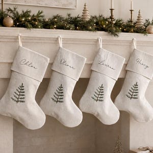 May include: Four off-white fabric Christmas stockings hanging in a row, each featuring a dark green embroidered pine tree design and a personalised name in black script: Chloe, Oliver, Lauren, and Henry, suspended by white fabric loops.
