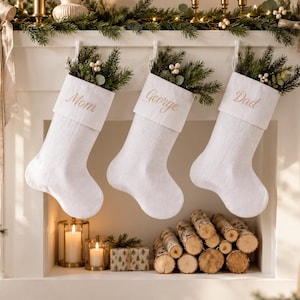 Peut inclure: Trois chaussettes de Noël en tissu blanc suspendues en rangée. Chaque chaussette présente un revers replié brodé d'un prénom en fil doré : Mom, George et Dad. Les chaussettes sont garnies de branches de pin vert et de petites baies blanches.