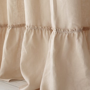 Puede incluir: Primer plano de una cortina de ducha de lino beige con un borde inferior con volantes. La cortina est&aacute; colgada en un ba&ntilde;o y el borde inferior descansa sobre un suelo de madera blanco.