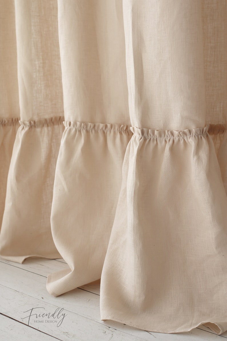 Puede incluir: Primer plano de una cortina de ducha de lino beige con un borde inferior con volantes. La cortina est&aacute; colgada en un ba&ntilde;o y el borde inferior descansa sobre un suelo de madera blanco.