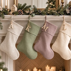 Peut inclure: Quatre chaussettes de Noël suspendues en rangée, présentant une palette de couleurs beige clair, vert sauge, mauve poussiéreux et crème. Chaque chaussette comprend un revers plié et une étiquette en bois avec les lettres A, K, M et L.