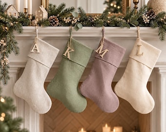 May include: Four Christmas stockings hanging in a row, featuring a light beige, sage green, dusty mauve, and cream color palette. Each stocking includes a folded cuff and a wooden letter tag attached to the top loop, displaying the letters A, K, M, and L.