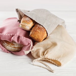 May include: Three loaves of bread in reusable cloth bags. The bags are pink, beige, and gray. The bread is partially visible in the bags.
