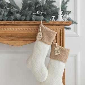 May include: Two white Christmas stockings with a tan cuff hang from a wooden mantel. Each stocking has a wooden tag with a letter. The mantel is decorated with greenery and a white candle.