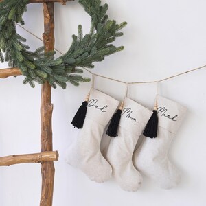 Puede incluir: Tres calcetines navide&ntilde;os de tela color hueso colgados de un cordel. Cada calcet&iacute;n tiene una borla negra, cuentas de madera y el texto en cursiva negra Dad, Mom y Mark. Una escalera de madera r&uacute;stica con una corona verde est&aacute; al lado.