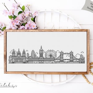 May include: A black and white line art illustration of the London skyline, including Big Ben, the London Eye, and Tower Bridge. The illustration is framed in a brown wooden frame and is displayed on a white surface.