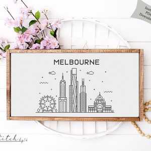 May include: A black and white line art illustration of the Melbourne skyline, including the Melbourne Star Observation Wheel, Federation Square, and other iconic buildings. The illustration is framed in a light brown wood frame and is set against a white background.