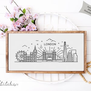 May include: Black and white line art cross stitch pattern of the London skyline, including the London Eye, Big Ben, Tower Bridge, and The Shard. The pattern is framed in a light brown wood frame and is displayed on a white surface. The text "LONDON" is included in the pattern.