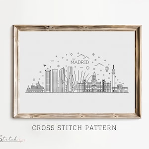 May include: Black and white line art cross stitch pattern of the Madrid skyline. The pattern includes the city's iconic buildings, including the Gran Vía, the Royal Palace, and the Temple of Debod. The pattern is framed in a light wood frame and includes the text "CROSS STITCH PATTERN" below the image.