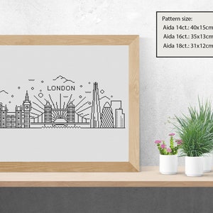 May include: A black and white line art illustration of the London skyline, including the London Eye, Big Ben, Tower Bridge, and The Shard. The illustration is framed in a light wood frame and is sitting on a wooden shelf with two potted plants.
