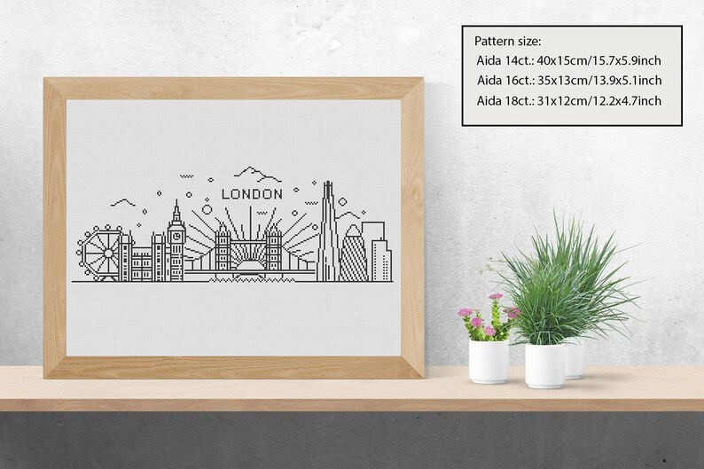 May include: A black and white line art illustration of the London skyline, including the London Eye, Big Ben, Tower Bridge, and The Shard. The illustration is framed in a light wood frame and is sitting on a wooden shelf with two potted plants.