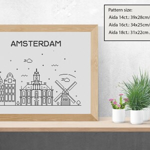 May include: A black and white line art illustration of the Amsterdam skyline, including buildings and a windmill, framed in a light wood frame. The text "AMSTERDAM" is at the top of the illustration. The image is on a wooden shelf with two potted plants.