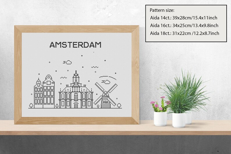 May include: A black and white line art illustration of the Amsterdam skyline, including buildings and a windmill, framed in a light wood frame. The text "AMSTERDAM" is at the top of the illustration. The image is on a wooden shelf with two potted plants.