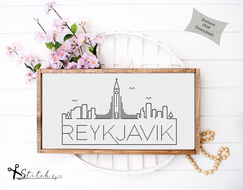 K&ouml;nnte beinhalten: Schwarzwei&szlig;es Kreuzstichmuster der Skyline von Reykjavik mit dem St&auml;dtenamen darunter. Das Muster ist in einem braunen Holzrahmen gerahmt und auf einer wei&szlig;en Oberfl&auml;che ausgestellt.