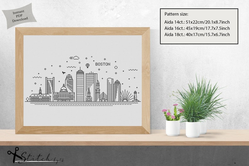 May include: A black and white line art illustration of the Boston skyline with the word "BOSTON" at the top. The illustration is framed in a light brown frame and is sitting on a wooden shelf with two potted plants.