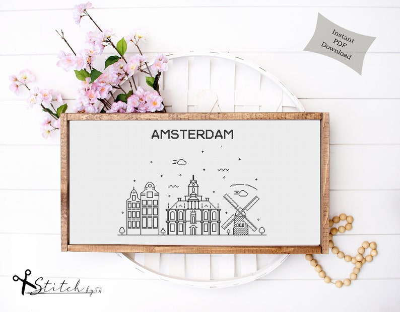 May include: A black and white line art illustration of the Amsterdam skyline, including a windmill, buildings, and the text "AMSTERDAM". The illustration is framed in a brown wood frame and displayed on a white surface.