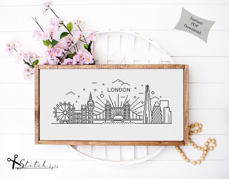 May include: Black and white line art cross stitch pattern of the London skyline, including the London Eye, Big Ben, Tower Bridge, and The Shard. The pattern is framed in a light brown wood frame and is displayed on a white surface. The text "LONDON" is included in the pattern.