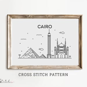 Może przedstawiać: Wzór haftu krzyżykowego w czerni i bieli przedstawiający panoramę Kairu z piramidami, meczetem i wysoką wieżą. Wzór jest oprawiony w jasną drewnianą ramę, a pod obrazem widnieje tekst "CAIRO" i "CROSS STITCH PATTERN".