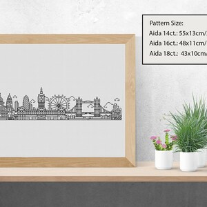 May include: A black and white line art illustration of the London skyline, including Big Ben, the London Eye, and Tower Bridge. The illustration is framed in a light wood frame and displayed on a shelf with potted plants.