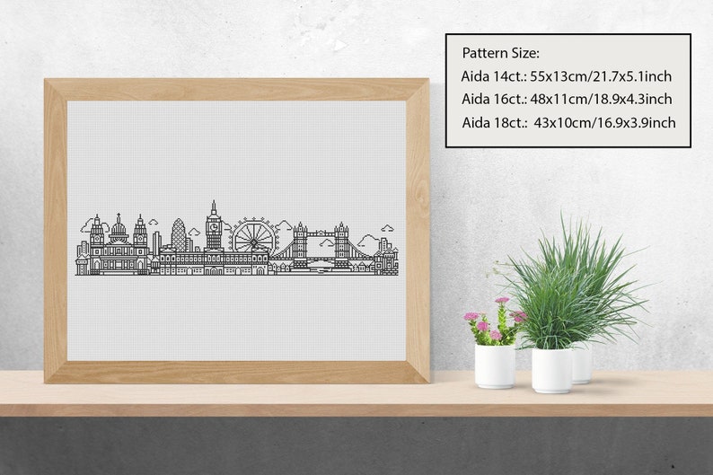 May include: A black and white line art illustration of the London skyline, including Big Ben, the London Eye, and Tower Bridge. The illustration is framed in a light wood frame and displayed on a shelf with potted plants.