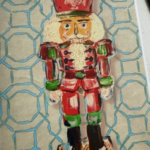 May include: A painting of a nutcracker figure wearing a red hat with a white fleur-de-lis emblem, a red and green uniform with black buttons, and black boots. The figure stands on a wooden base against a beige background with a blue geometric pattern.