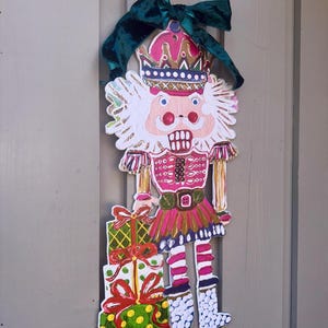 May include: A wooden Christmas nutcracker decoration with a pink and white face, wearing a pink and gold uniform, and holding wrapped gifts. The decoration is adorned with a green velvet bow at the top.