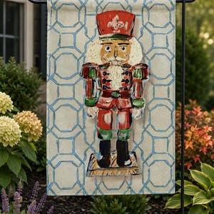 May include: A rectangular garden flag featuring a central nutcracker illustration in red, green, and black, set against a light beige background with a repeating blue geometric lattice pattern. The nutcracker wears a red hat with a white fleur-de-lis.