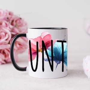 May include: White ceramic mug with a black handle and rim. The mug has a watercolor design of pink and blue flowers and the word "UNT" in black lettering.