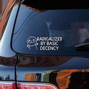 May include: A white vinyl car decal with the text "RADICALIZED BY BASIC DECENCY" and a simple cartoon figure. The decal is affixed to the rear window of a black vehicle, with the taillight visible.