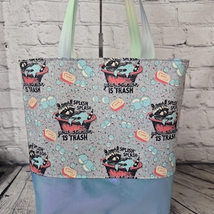 May include: A gray tote bag with a raccoon design. The raccoon is in a tub with bubbles and the text "your opinion is trash" is repeated on the bag. The bag has a light blue iridescent bottom.