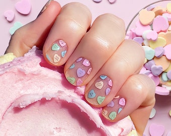 May include: Close-up of a hand with pastel heart-shaped nail art. The nails feature small, colorful hearts with phrases like "True Love" and "Be Mine." Scattered heart candies and a pink dessert are in the background.