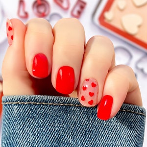 May include: Close-up of a hand with red nail polish.  Four nails have a solid red polish, and one nail has a clear coat with red hearts.