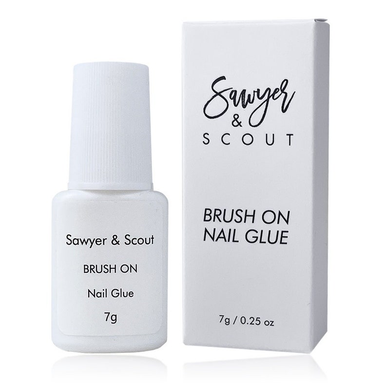 May include: A white bottle of Sawyer & Scout brush-on nail glue with a white label that says "Brush On Nail Glue" and "7g". The bottle is next to a white box with the same text and the additional text "7g / 0.25 oz".