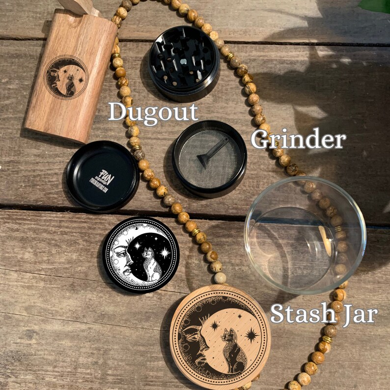 Puede incluir: Un conjunto de accesorios para fumar que incluye un dugout de madera, un grinder de metal, un tarro de almacenamiento de vidrio y dos tapas de madera con un dise&ntilde;o de luna y gato. Los accesorios est&aacute;n dispuestos sobre una superficie de madera con un collar de cuentas.