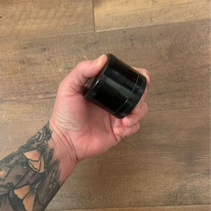 Puede incluir: Un molinillo de metal negro con una superficie texturizada. El molinillo est&aacute; hecho de tres piezas y tiene un peque&ntilde;o compartimento para recoger el kief.