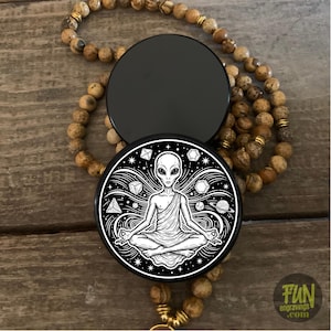 May include: A black and white graphic of an alien meditating, surrounded by geometric shapes and planets, on a round black object. A beaded necklace with brown and gold beads is draped around the object.