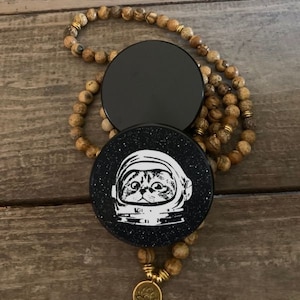 May include: A black and white phone grip with a cat wearing an astronaut helmet in space. The grip is attached to a brown and gold beaded necklace.