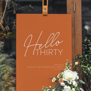 Puede incluir: Un cartel de bienvenida marrón con texto blanco que dice "Hello Thirty" y "Bienvenido a la fiesta de cumpleaños de Emilia por sus 30 años 12.19.2022". El cartel está en un caballete frente a una puerta de madera.
